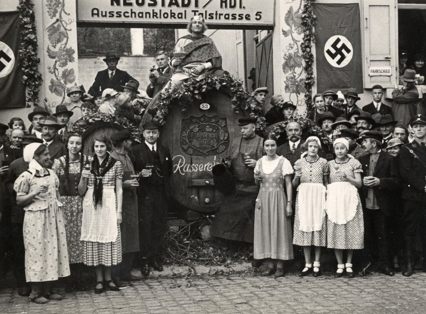 Vor einem großen Weinfass stehen Frauen in Schürzen und Männer mit gefüllten Weingläsern. Daneben hängt eine Hakenkreuzfahne.