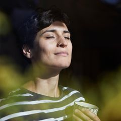 Eine Frau hat die Augen geschlossenen und steht mit einer Tasse Tee am Fenster