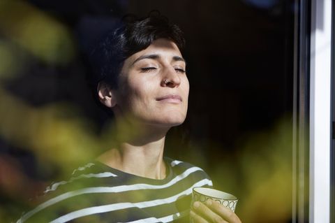 Eine Frau hat die Augen geschlossenen und steht mit einer Tasse Tee am Fenster