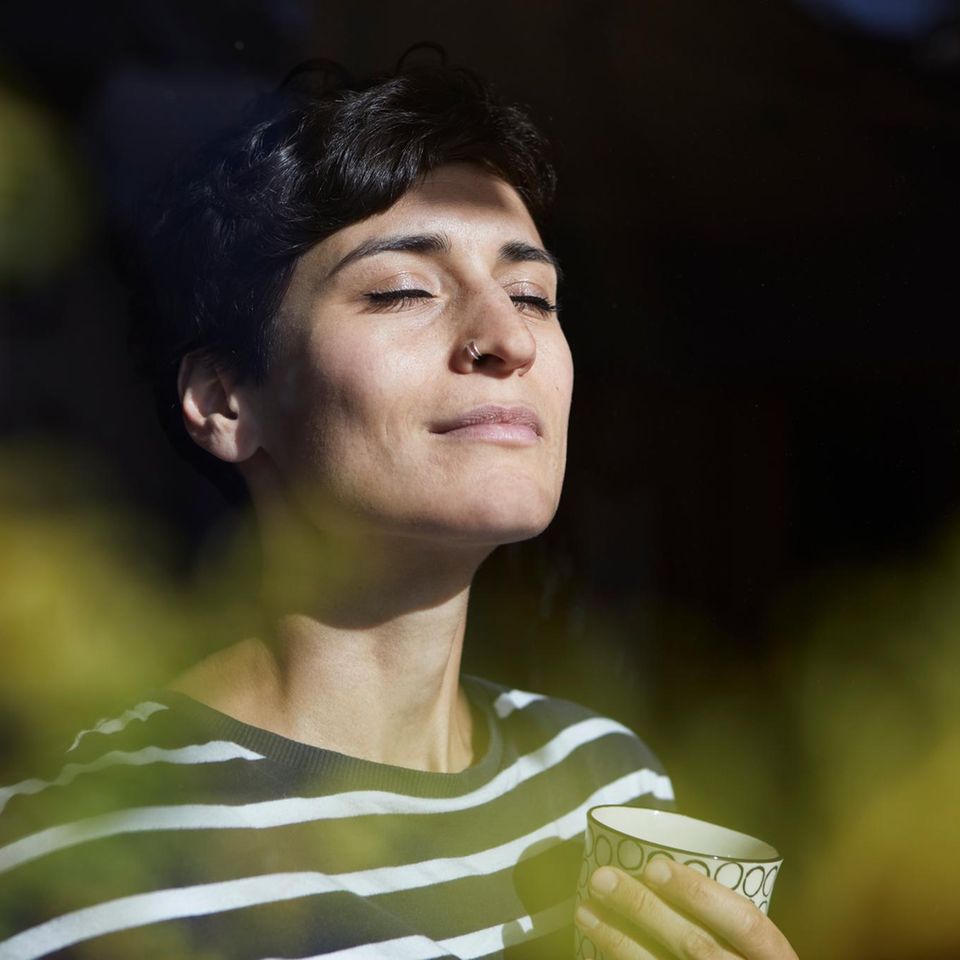 Eine Frau hat die Augen geschlossenen und steht mit einer Tasse Tee am Fenster