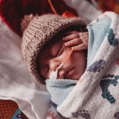 Ein Frühgeborenes in einer Decke eingewickelt hält eine Hand an seinem Ohr