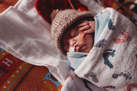 Ein Frühgeborenes in einer Decke eingewickelt hält eine Hand an seinem Ohr Ein Frühgeborenes in einer Decke eingewickelt hält eine Hand an seinem Ohr