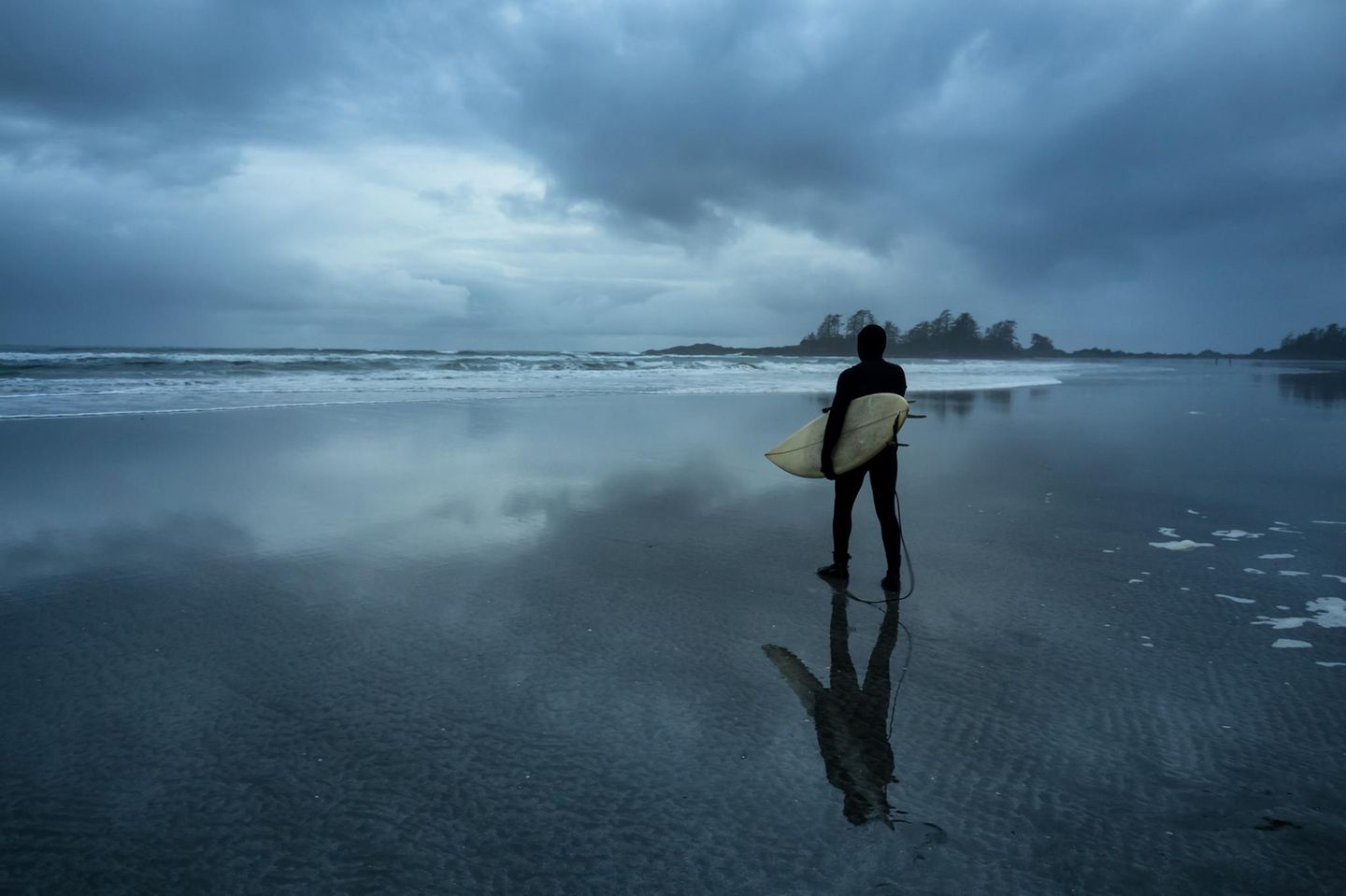 Surfer auf dem Weg in den Ozean während eines bewölkten Wintersonnenuntergangs. Aufgenommen in Chesterman Beach, Tofino, Vancouver Island, BC, Kanada.