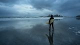 Surfer auf dem Weg in den Ozean während eines bewölkten Wintersonnenuntergangs. Aufgenommen in Chesterman Beach, Tofino, Vancouver Island, BC, Kanada.
