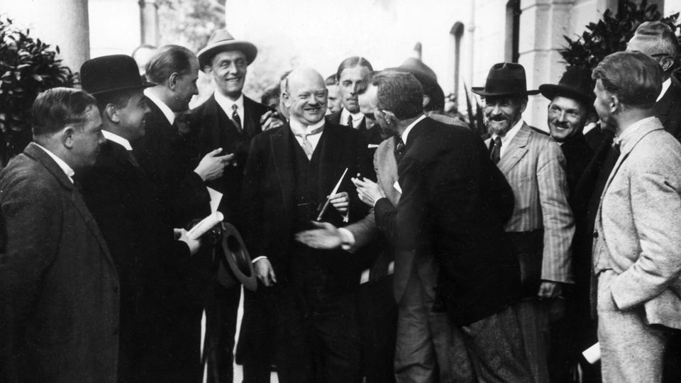 Ein lachender Gustav Stresemann geht durch eine Gruppe von Journalisten