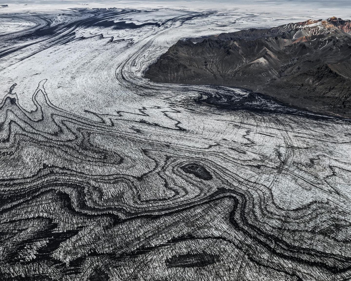 Wenn sich Gletscherströme ihren Weg durch das Gebirge bahnen, verschlägt ihre schiere Kraft den Betrachtenden meist den Atem. Die schwarze Vulkanasche, die das Eis des Skeidararjökull hier marmoriert, zeugt jedoch von einem noch gewaltigeren Naturspektakel: dem Wechselspiel zwischen Schnee und Feuer, Sonne und Wind. "Die Vulkaneruptionen liefern die schwarze Asche, welche die Gletscher überdeckt. Darauf fällt frischer Schnee, der allmählich zu Eis komprimiert wird. So legt sich weiße auf schwarze Schicht, viele Male", beschreibt die Geologin Angelika Jung-Hüttl das Phänomen. Je weiter sich der Gletscher seinen Weg ins Tal bahnt, desto verwobener sind die beiden Schichten, desto schwungvoller die Muster.