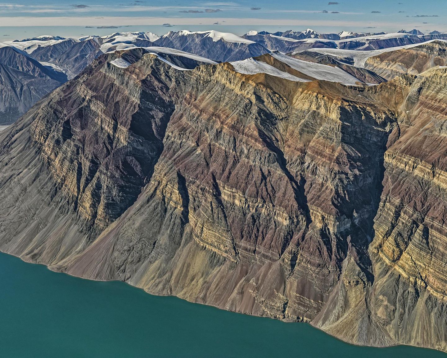 Wie bei einer Schichttorte haben sich die unterschiedlich gefärbten Gesteinsschichten im Laufe der Jahrmillionen zu dem Gebirgsstock gestapelt, der heute am Rande des Kong-Oscar-Fjords in Ostgrönland thront. Der schroffe Fels überragt den Fjord um 1900 Meter und zeugt noch heute von seiner einstigen Lage am Rande eines urzeitlichen Ozeans. Auf dessen Grund lagerten sich in verschiedenen geologischen Zeiten verschiedenfarbige Materialien ab. Dass diese Schichtung noch heute zu bewundern ist, ist der Tatsache zu verdanken, dass das Gebirge während der Kaledonischen Gebirgsbildung vor 400 Millionen Jahren – im Gegensatz zu anderen Bergzügen – nicht gefaltet, sondern lediglich angehoben, leicht gekippt und in Blöcke zerlegt wurde.