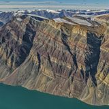 Wie bei einer Schichttorte haben sich die unterschiedlich gefärbten Gesteinsschichten im Laufe der Jahrmillionen zu dem Gebirgsstock gestapelt, der heute am Rande des Kong-Oscar-Fjords in Ostgrönland thront. Der schroffe Fels überragt den Fjord um 1900 Meter und zeugt noch heute von seiner einstigen Lage am Rande eines urzeitlichen Ozeans. Auf dessen Grund lagerten sich in verschiedenen geologischen Zeiten verschiedenfarbige Materialien ab. Dass diese Schichtung noch heute zu bewundern ist, ist der Tatsache zu verdanken, dass das Gebirge während der Kaledonischen Gebirgsbildung vor 400 Millionen Jahren – im Gegensatz zu anderen Bergzügen – nicht gefaltet, sondern lediglich angehoben, leicht gekippt und in Blöcke zerlegt wurde.