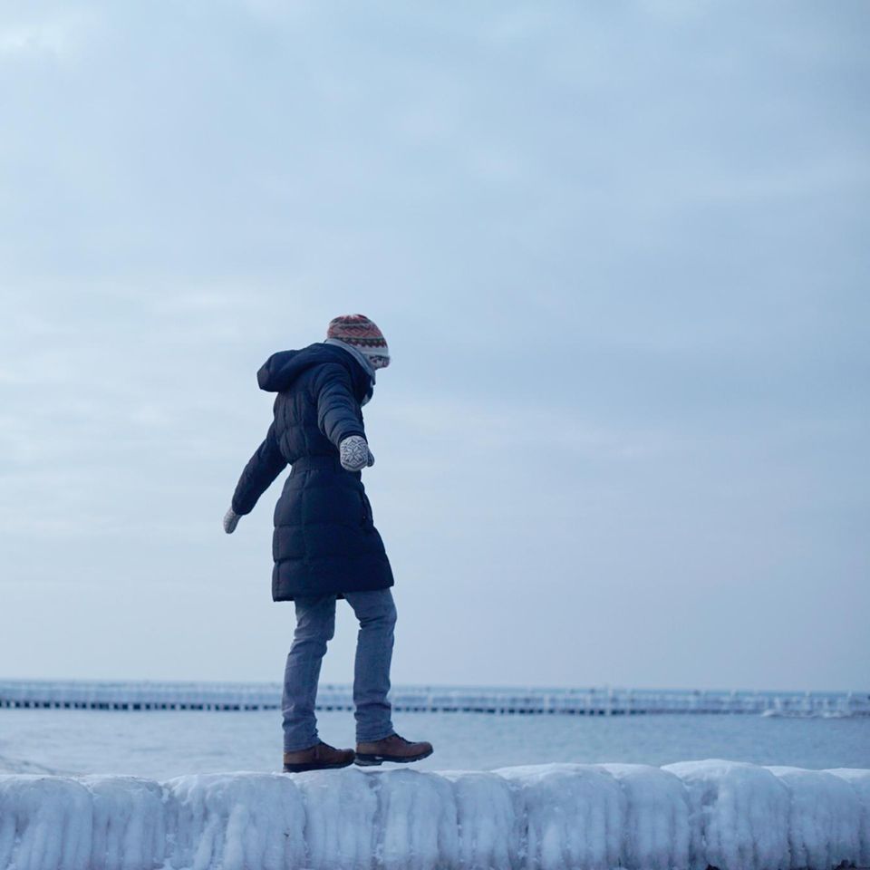 Frau balanciert unsicher auf einer glatten Buhne aus Eis am Meer
