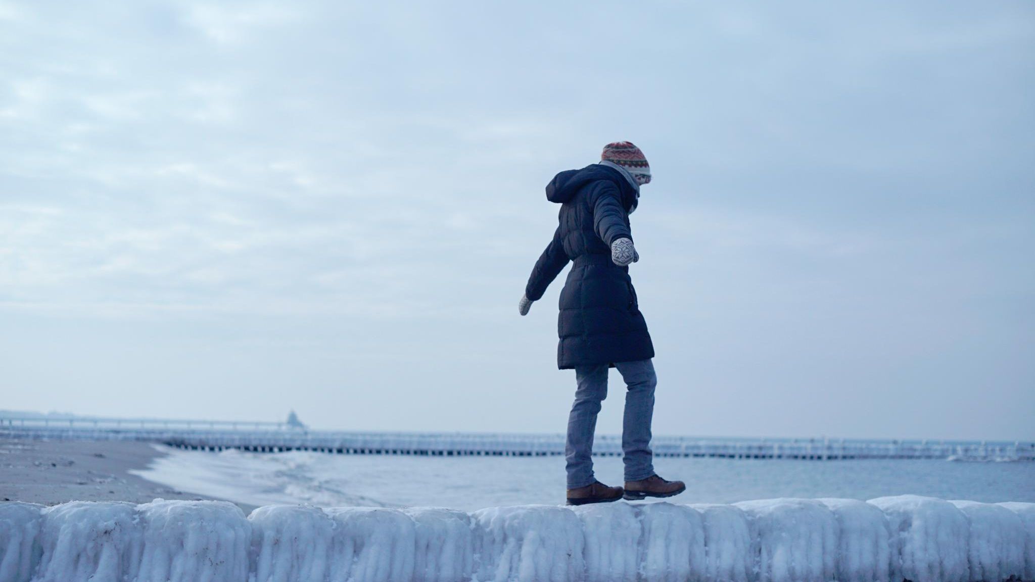 Frau balanciert unsicher auf einer glatten Buhne aus Eis am Meer