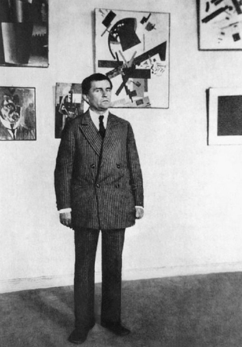 Kasimir Malewitsch posiert vor einer Wand mit Gemälden