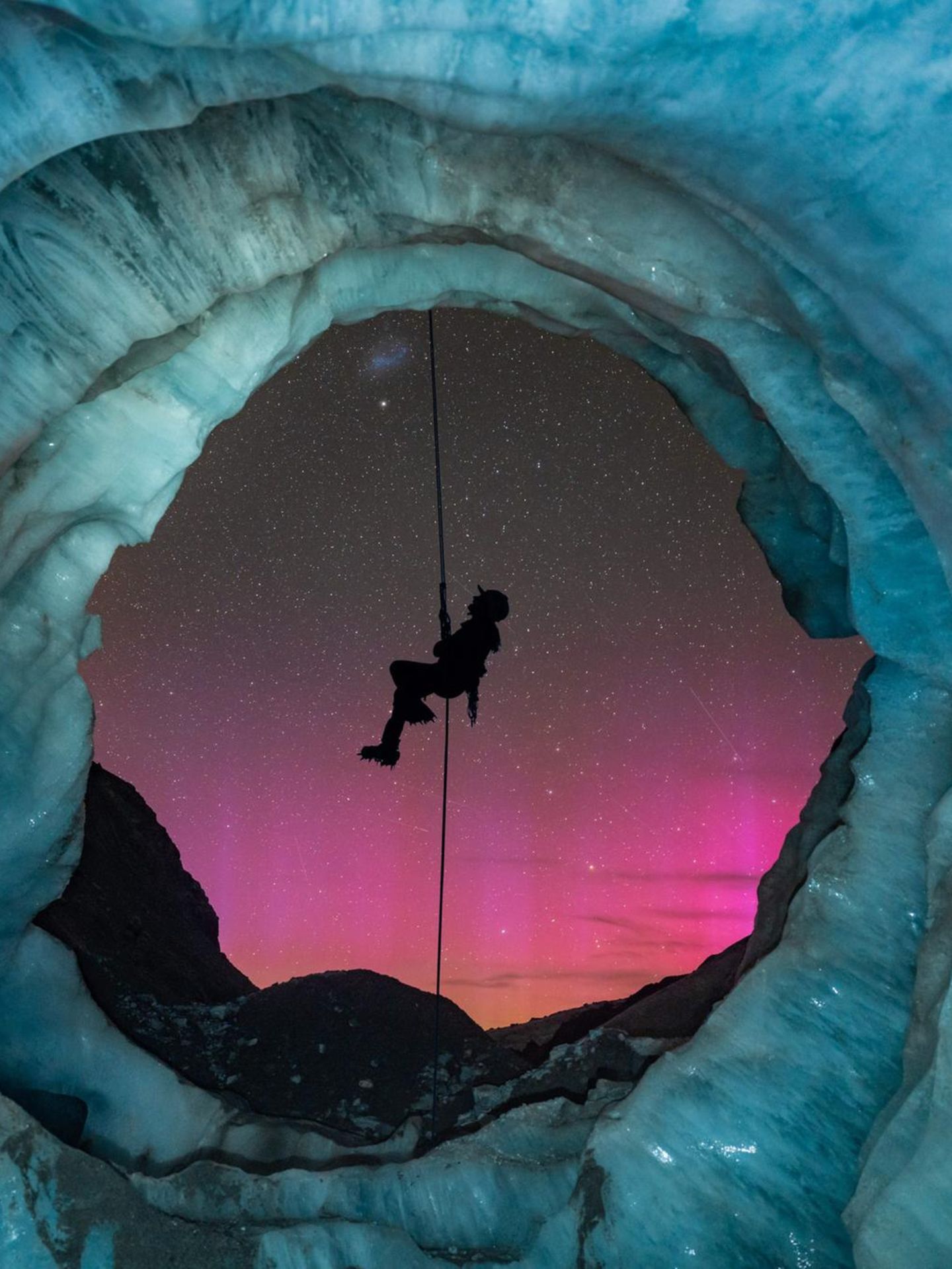 Blick aus einer blauen Eishöhle auf einen rosafarbenen Sternenhimmel, davor die Silhouette eines Bergsteigers am Seil