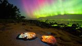 Grün-violettes Polarlicht über einem Strand; darauf leuchtende Steine