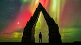Ein dunkler spitz zulaufender Steinbogen, darin die Silhouette eines Menschen, darüber Polarlichter in gelb, rot und grün
