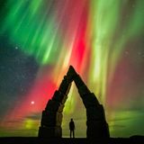 Ein dunkler spitz zulaufender Steinbogen, darin die Silhouette eines Menschen, darüber Polarlichter in gelb, rot und grün