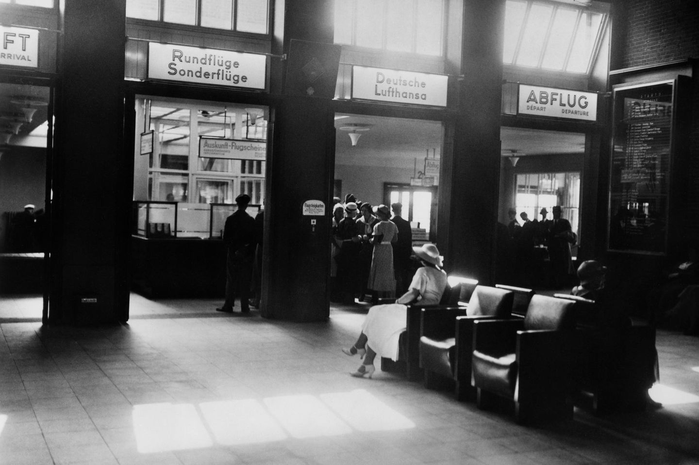 Eine Abfertigungshalle der Lufthansa mit Sesseln und Menschen vor Schaltern, schwarz-weiß Foto