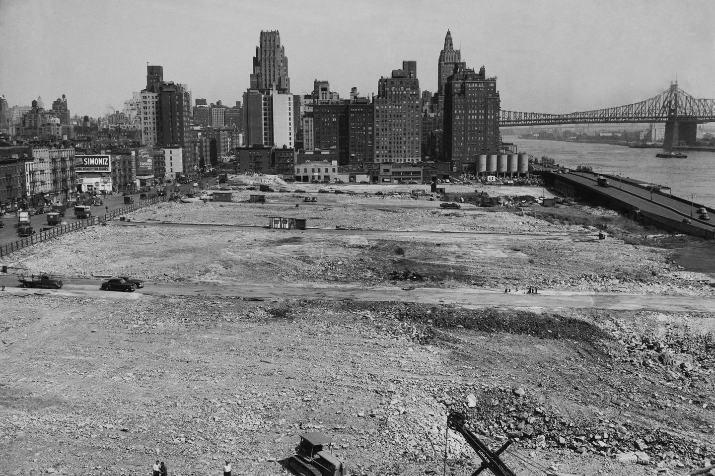 Schwarz-weiß Aufnahme eines Baugrundstücks an einem Fluss, im Hintergrund Hochhäuser und eine Brücke
