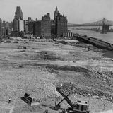 Schwarz-weiß Aufnahme eines Baugrundstücks an einem Fluss, im Hintergrund Hochhäuser und eine Brücke