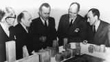 Männer in dunklen Anzügen vor einem Architekturmodell aus groben Quadern auf einem weißen Tisch, sw-Foto