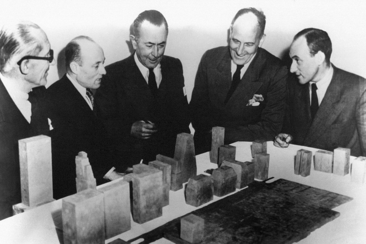 Männer in dunklen Anzügen vor einem Architekturmodell aus groben Quadern auf einem weißen Tisch, sw-Foto