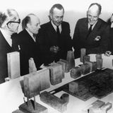 Männer in dunklen Anzügen vor einem Architekturmodell aus groben Quadern auf einem weißen Tisch, sw-Foto