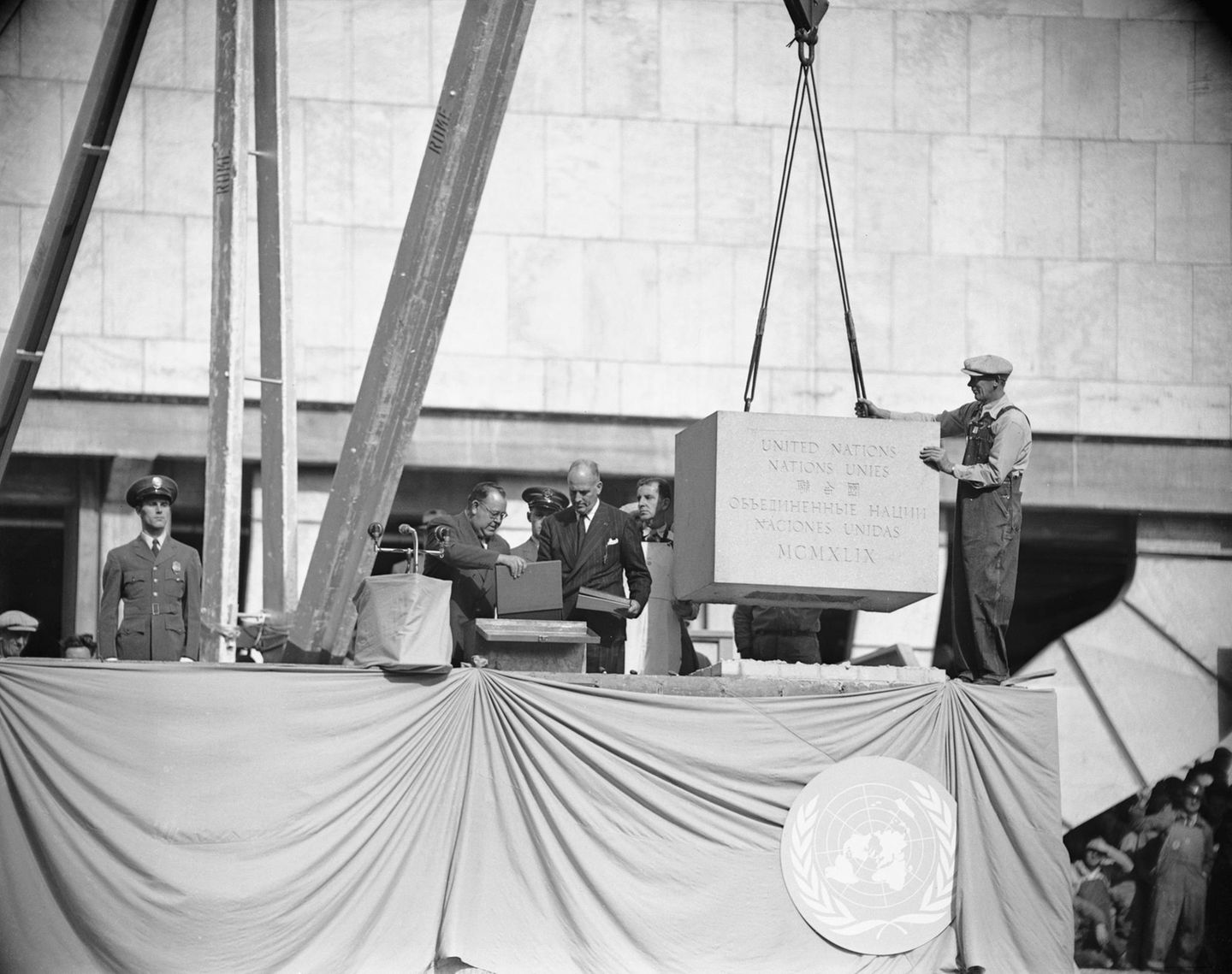 Ein gewaltiger Grundstein wird von einem Arbeiter in Position bugsiert; daneben Offizielle, sw-Foto 1949