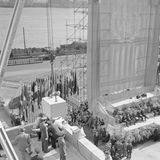 Festgesellschaft auf einer Tribüne beobachtet das Asenken eines Grundsteins, dahinter die Flagge der Vereinten Nationen, sw-Foto