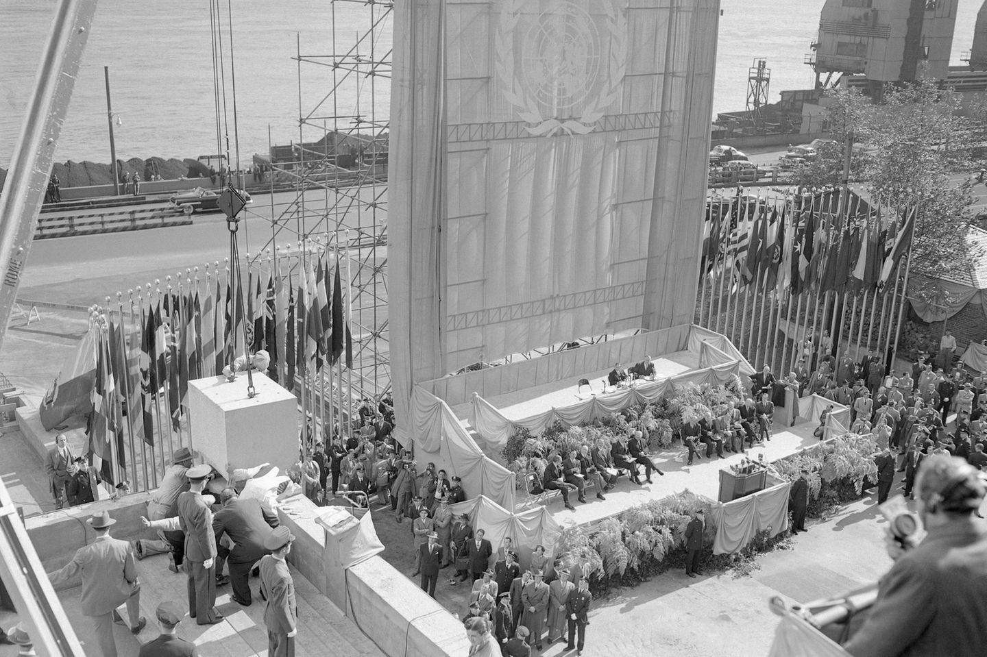 Festgesellschaft auf einer Tribüne beobachtet das Asenken eines Grundsteins, dahinter die Flagge der Vereinten Nationen, sw-Foto