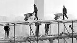 Arbeiter mit Schubkarren auf einem Baugerüst, dahinter die Silhouette eines Wolkenkratzers; sw-Foto, 1949
