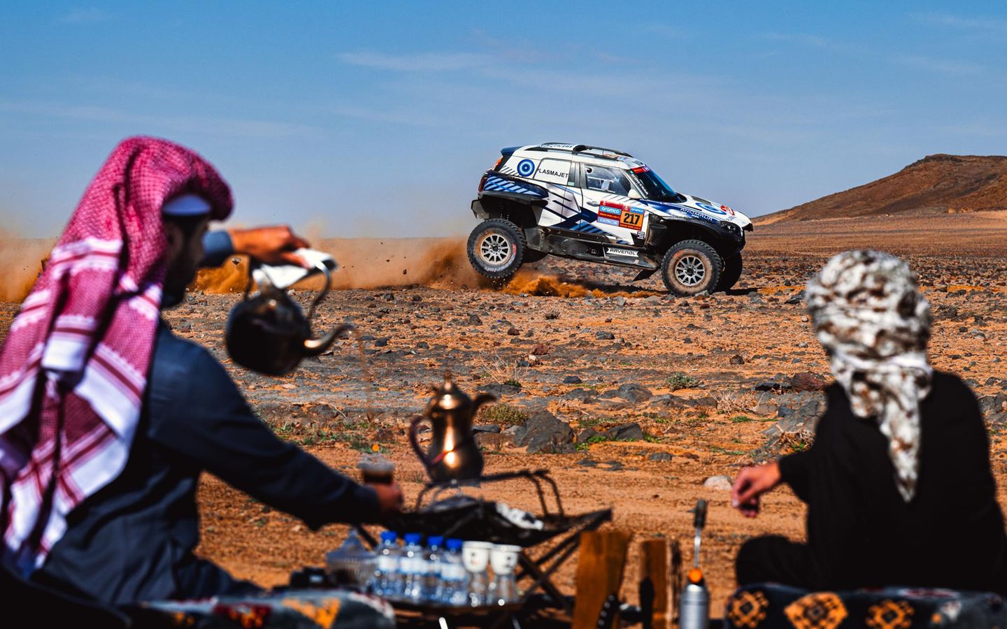 Zuschauer bereiten in Saudi-Arabien auf traditionelle Weise Tee zu, während im Hintergrund ein Rennwagen der Rallye Dakar Staub aufwirbelt. Der Wettbewerb hat eine lange Geschichte, hieß ursprünglich "Ralley Paris-Dakar", gastierte auch schon in Südamerika und wird inzwischen von Saudi-Arabien ausgerichtet. Wegen vieler Todesfälle, hoher Kosten und ökologischer Folgen steht die Veranstaltung seit Jahren in der Kritik. 