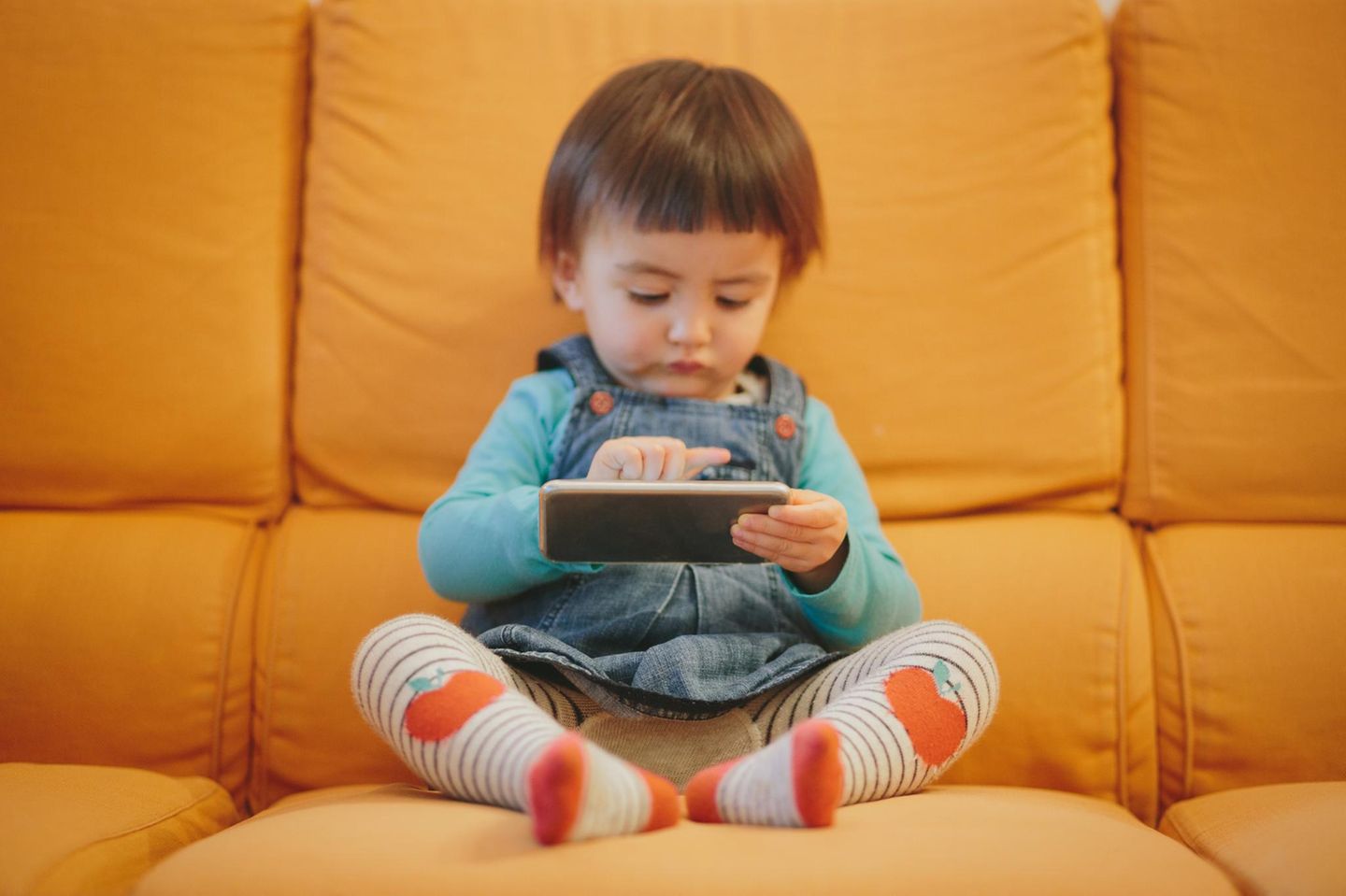 Ein Kleinkind sitzt auf der Couch und spielt mit einem Smartphone