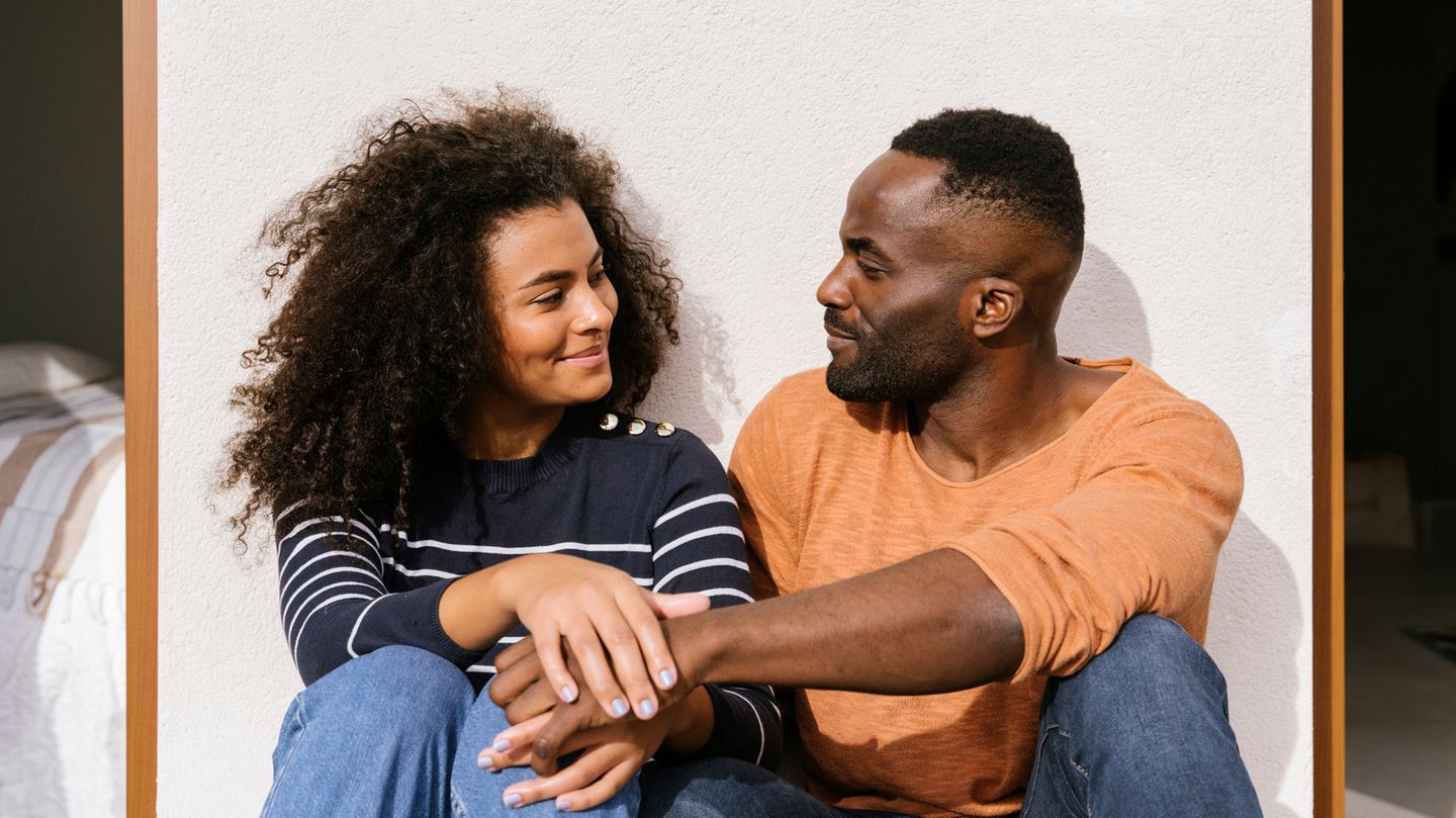 Ein Paar hockt vor einer Wand, lächelt sich an und hält einander die Hand
