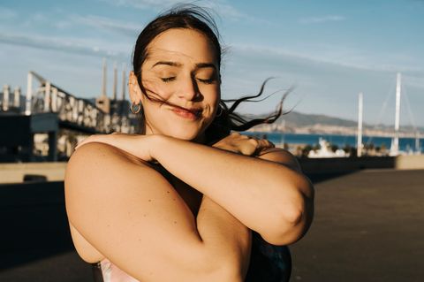 Eine Person umarmt sich selbst und steht mit geschlossenen Augen im Sonnenlicht