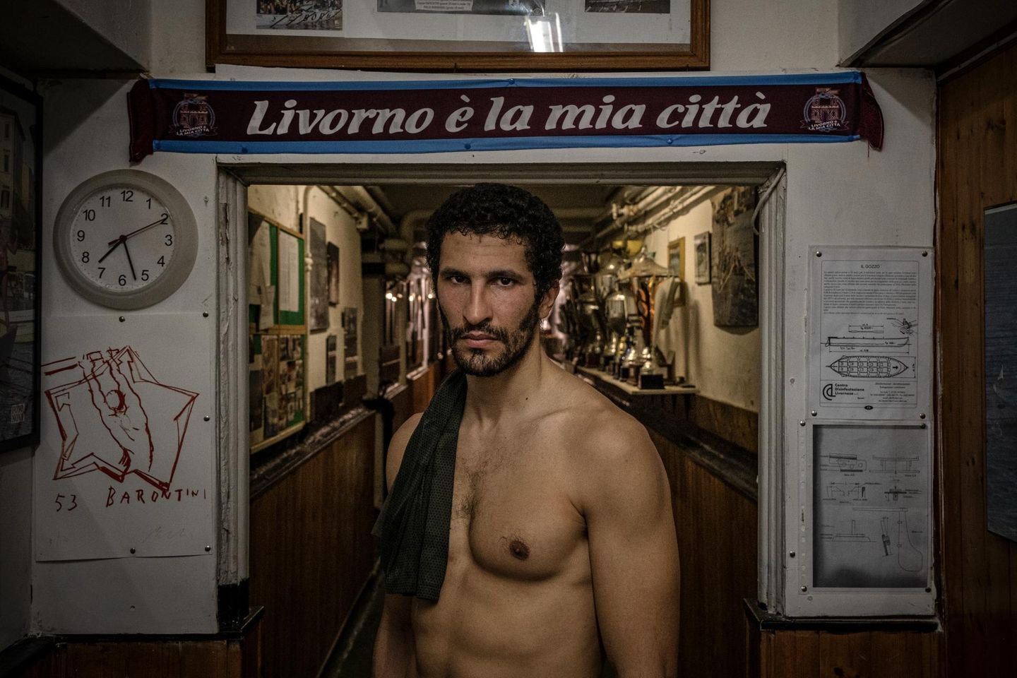 Im historischen Stadtteil Borgo Cappuccini im italienischen Livorno steht Karim Rachedi vor dem Eingang eines lokalen Clubs. Der Blick des Athleten wirkt ruhig. Und  dabei auch ein Stückweit herausfordernd. "Livorno ist meine Stadt" prangt auf dem Schal hinter dem Ruderer; ein kurzer Satz, der sportliche Disziplin mit Zugehörigkeit verknüpft. Denn das Rudern fordert hier nicht bloß Wettkampfsgeist. Es prägt auch Identität: ein kollektives Ritual zwischen Hafen und Meer, Wohnhäusern und Vereinsraum. Die Pokale im Hintergrund erzählen von Siegen, Jubel, Tradition. 