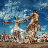 "Die Füße im Matsch, die Herzen im Göttlichen" lautet der ursprüngliche Titel dieses Fotos, das vor Dynamik nur so strotzt. Zwei Männer tanzen mit sichtlicher Freude und messen sich in einem symbolischen Stockkampf. Über der Schulter tragen sie die traditionellen langen Bambusstöcke mit orangefarbenen Fahnen, die im Wind spielen. Ihr Pulikum jubelt und klatscht. Zuschauer und Tanzende gehören zu den "Varkari", einer indischen Religionskultur, deren Anhänger großen Wert auf Gesang und Musik legen. 