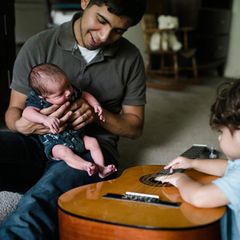 Vater hält Neugeborenes auf dem Schoss während der größere Bruder auf einer Gitarre musiziert.