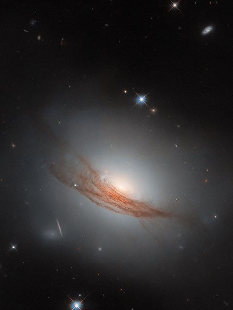 NGC 7722 ist ein kosmisches Zwischenwesen, das Eigenschaften von Spiralgalaxien und elliptischen Galaxien vereint – und doch keiner dieser Gruppen angehört. Forscher zählen sie zu der kleinen, schwer definierbaren Gruppe der linsenförmigen Galaxien. Rund 187 Millionen Lichtjahre von der Erde entfernt erstrahlt die kosmische Schönheit im luftleeren Raum. Sie besitzt den leuchtenden Halo einer elliptischen Galaxie und eine Scheibe, um die konzentrische Staubringe wirbeln. Ihr charakteristischstes Merkmal ist der rostrote Schleier: Schwaden aus Staub winden sich um die leuchtende Mitte der Sterneninsel. Viele linsenförmige Galaxien tragen solch ein diffuses Band. Forschende sehen darin Spuren der Verschmelzung mit einer anderen Galaxie.   2020 verfolgten Astronomen und Astrophysikerinnen gebannt, wie ein Stern in den Sphären von NGC 7722 explodierte. Vorübergehend überstrahlte die Supernova ihre kosmische Nachbarschaft; inzwischen ist sie jedoch verglüht. So gelang es dem Hubble-Weltraumteleskop, diese außergewöhnliche Sterneninsel schärfer einzufangen als je zuvor.  KW06