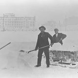 Drei Männer mit Schaufeln schauen in die Kamera, sie schippen Schnee in einen großen Trog auf einem Schlitten