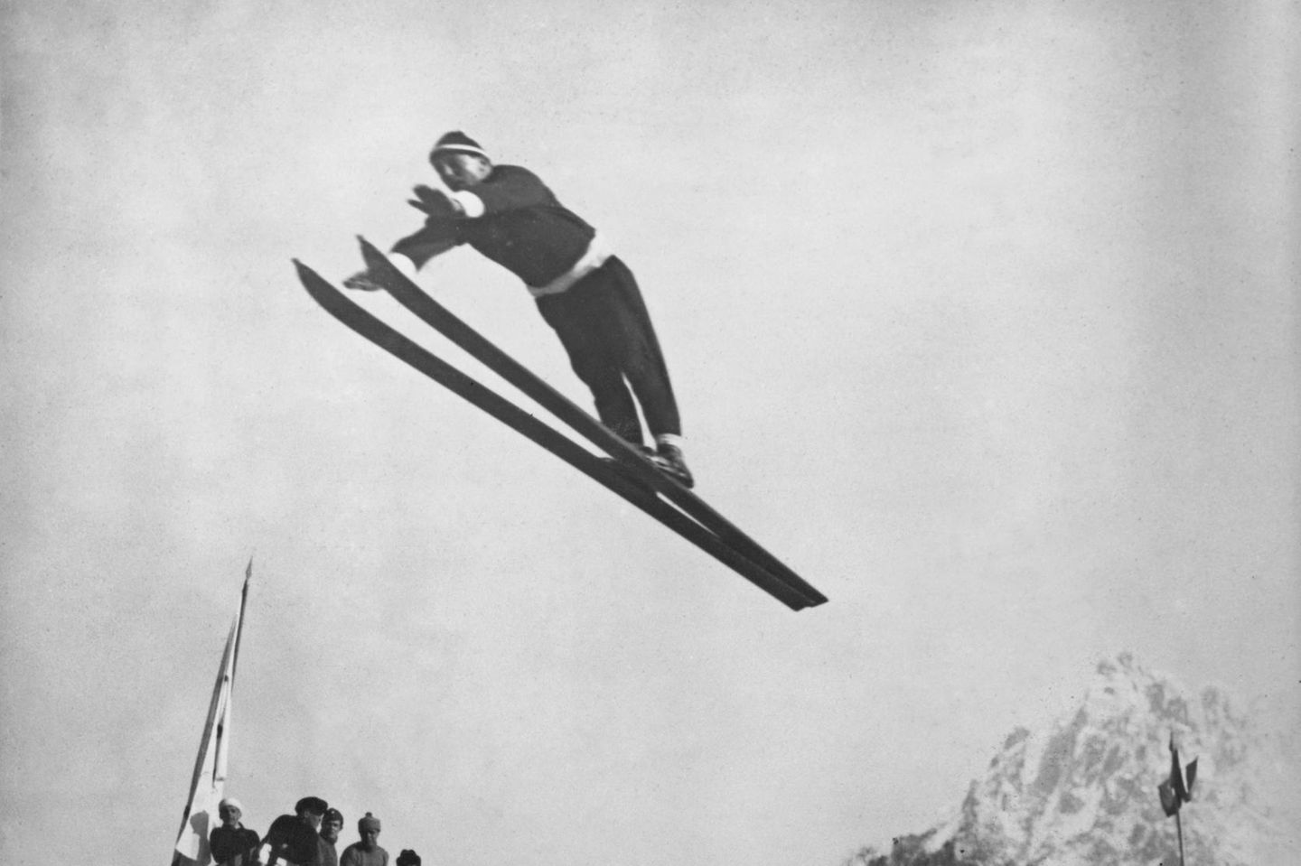 Ein Skispringer mit Pudelmütze in der Luft, Skier zusammengehalten, Arme nach vorne gestreckt.