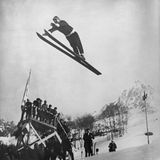 Ein Skispringer mit Pudelmütze in der Luft, Skier zusammengehalten, Arme nach vorne gestreckt.