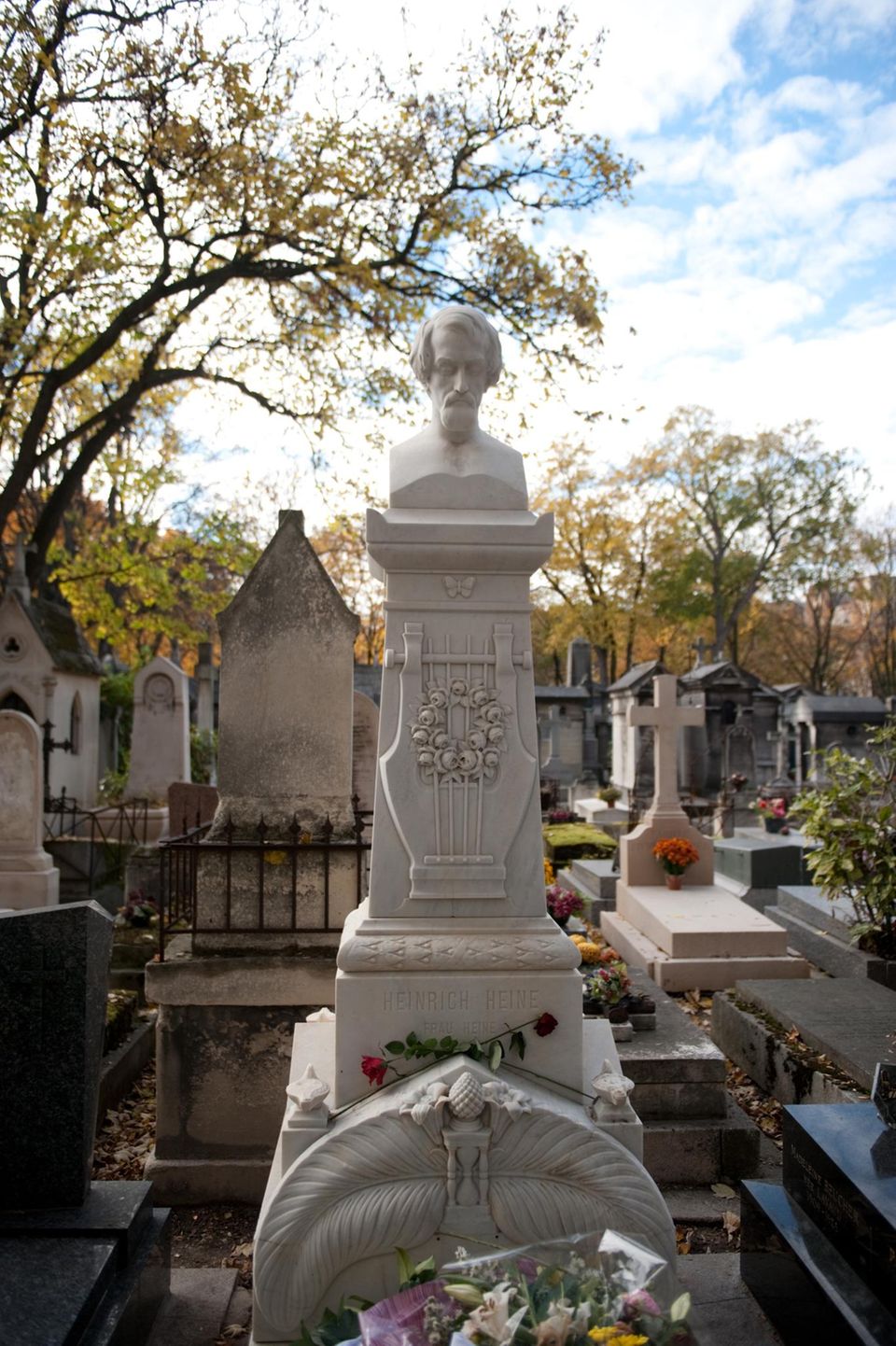 Auf einem Friedhof steht ein weißes Grabmal, bekrönt von einem männlichen Kopf.