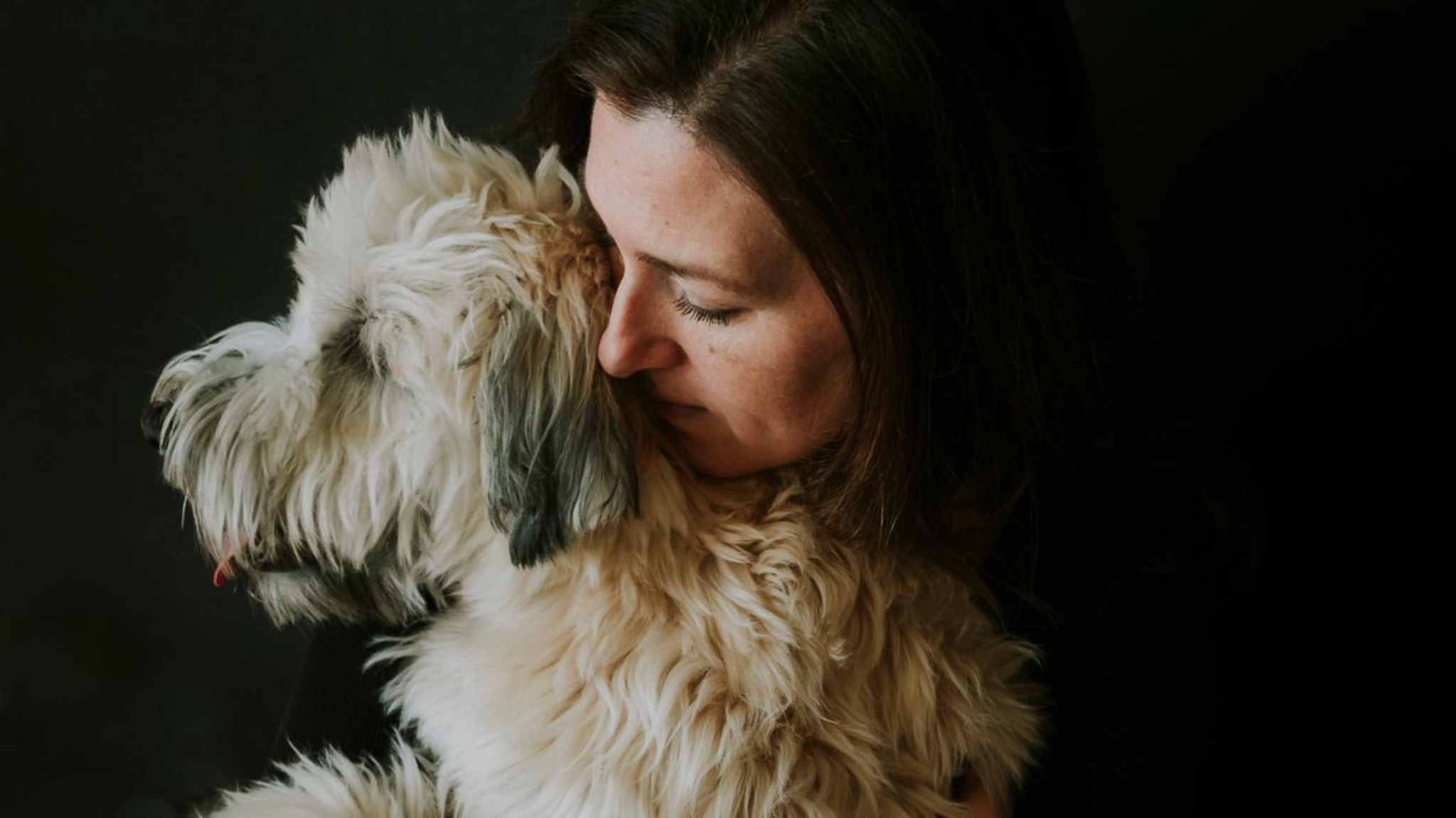 Frau umarmt weissen flauschigen Hund vor schwarzem Hintergrund