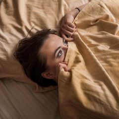 Eine Frau liegt im Bett, die Bettdecke halb über ihr Gesicht gezogen