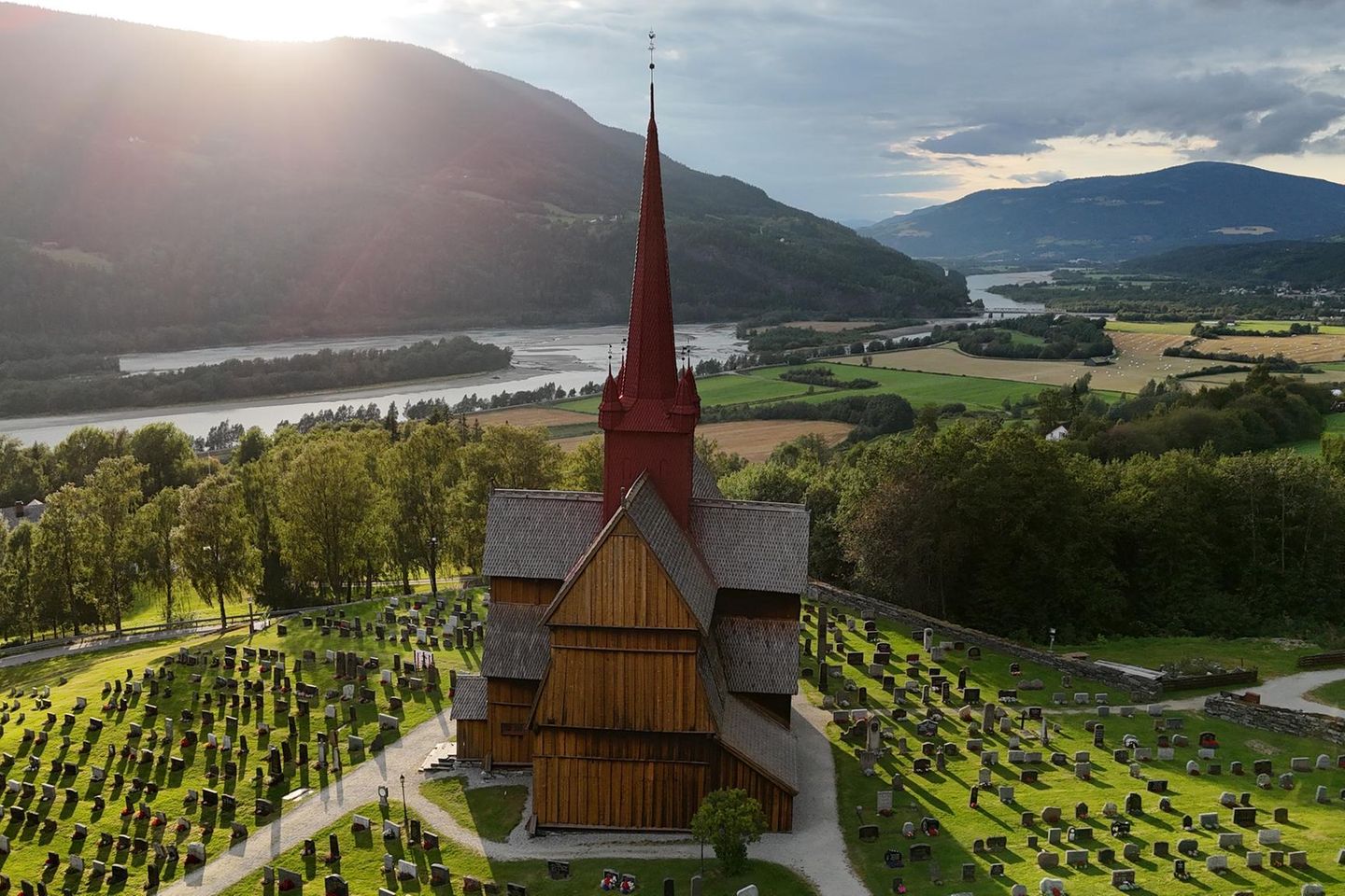 Ringebu Stabkirche, umringt von Bäumen und Feldern, im Hintergrund Berge