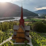 Ringebu Stabkirche, umringt von Bäumen und Feldern, im Hintergrund Berge