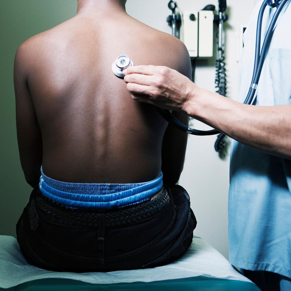 Ein weisshäutiger Arzt hört den Rücken einer dunkelhäutigen Person ab
