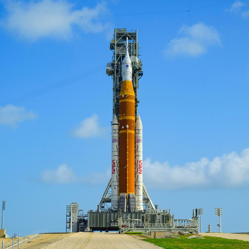 Auf der Startrampe in Florida steht die Rakete schon bereit. An ihrer Spitze ist die Orionkapsel (weiß) zu sehen, in der die Crew sitzen wird. Darunter das Space Launch System (orange, mit weißen "Boostern"), das die Kapsel von der Erde tragen und auf ihre Reise Richtung Mond schubsen soll