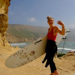 Frau mit Surfboard vor bergiger Kulisse und Meer