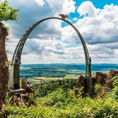 Bogen mit Adlerfigur vor weiter Landschaft und wolkigem Himmel