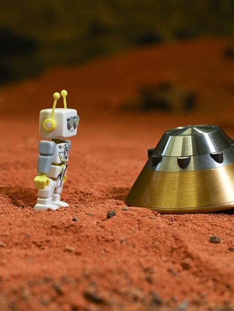 Ein Spielzeug-Marsmensch steht auf rotem Untergrund und blickt auf eine  Testkapsel des ExoMars-Landemoduls.